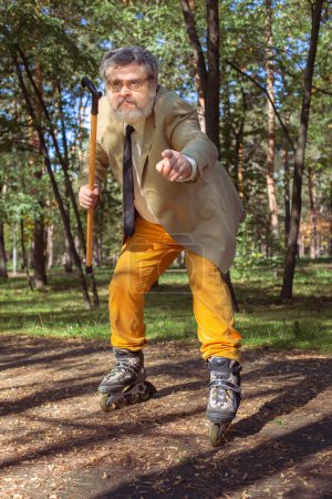 Foto de El viejo gracioso patina en el parque. Un abuelo activo con un bastón baila y hace caras. Abuelo con barba blanca. - Imagen libre de derechos