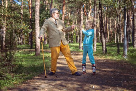 Téléchargez les photos : Grand-père marche avec sa petite-fille dans le parc. Une fille patine à roulettes dans le parc avec son vieux grand-père. Un grand-père drôle et une petite fille jouent et plaisantent. - en image libre de droit
