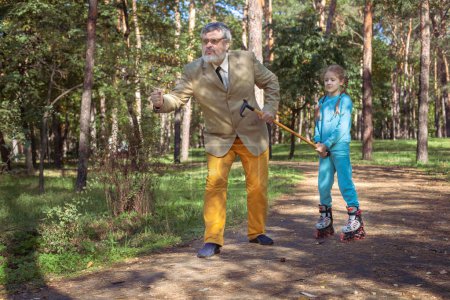 Foto de El abuelo camina con su nieta en el parque. Una chica patina en el parque con su abuelo. Un abuelo divertido y una niña están jugando y bromeando. - Imagen libre de derechos