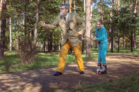 Téléchargez les photos : Grand-père marche avec sa petite-fille dans le parc. Une fille patine à roulettes dans le parc avec son vieux grand-père. Un grand-père drôle et une petite fille jouent et plaisantent. - en image libre de droit