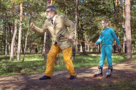 Photo for Grandfather walks with his granddaughter in the park. A girl roller skates in the park with her old grandfather. A funny grandfather and a little girl are playing and joking. - Royalty Free Image