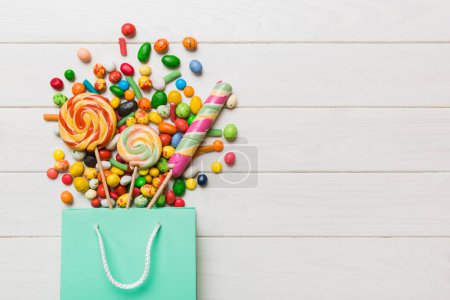 Téléchargez les photos : Shopping paper gift bag in corner full of assorted traditional candies falling out on colored background with copy space. Happy Holidays sale concept. - en image libre de droit