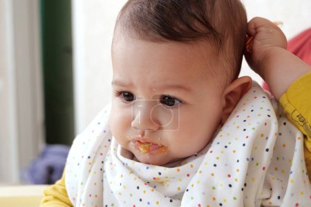 Portrait of little baby boy eating food. Baby with a spoon in feeding chair. Cute baby eating first meal.