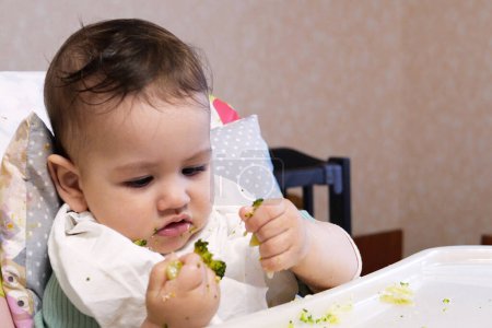 Portrait of little baby boy eating food. Baby with a spoon in feeding chair. Cute baby eating first meal.