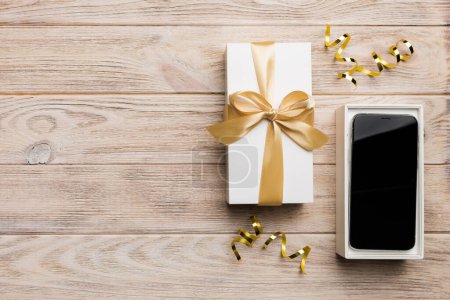 Geöffnete Geschenkbox mit Goldband und Smartphone auf farbigem Hintergrund, Ansicht von oben. Offene Verpackungsattrappe, Vorlage für Ihr Design - Branding-Attrappe.