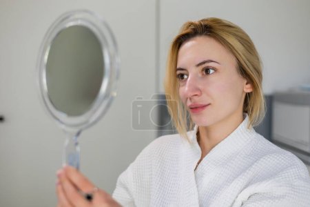 Nice beautiful girl with natural beauty, holding a mirror in hand, dressed in a white robe, looks at herself after beauty procedures, such as carbon peeling and skin rejuvenation, in a beauty clinic