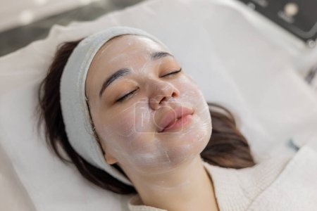 Photo for Stunning brunette woman with a white cream mask on her face reclines in a beauty and wellness clinic. Girl luxuriates in skin rejuvenation and care treatments at a spa salon, light smile on face - Royalty Free Image