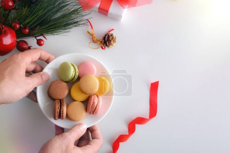 Téléchargez les photos : Mains servant pile de macarons français de différentes saveurs dans une assiette en céramique sur table blanche avec décoration de Noël. Vue du dessus. - en image libre de droit