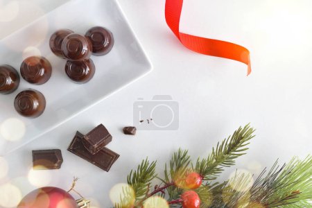 Téléchargez les photos : Assiette en céramique blanche remplie de bonbons au chocolat noir sur une table blanche avec des décorations de Noël et des portions de choclolate pour desserts. Vue du dessus. - en image libre de droit