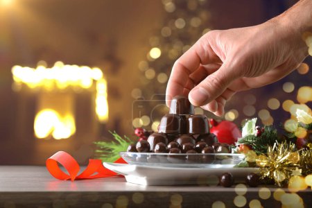 Téléchargez les photos : Ramasser à la main un bonbon au chocolat noir à partir d'une pile de bonbons au chocolat sur une table en bois avec décoration de Noël et fond chaud de fête à la maison. Vue de face. - en image libre de droit