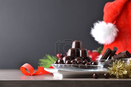 Téléchargez les photos : Pile de bonbons au chocolat empilés dans une plaque de verre avec arc rouge et décoration de Noël sur fond sombre isolé. Vue de face. - en image libre de droit