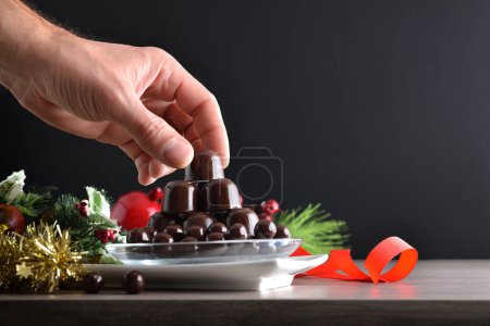 Téléchargez les photos : Ramasser à la main un bonbon au chocolat dans une assiette sur une table en bois avec décoration de Noël et fond sombre isolé. Vue de face. - en image libre de droit
