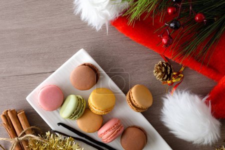 Téléchargez les photos : Empilement de différentes saveurs macarons français servis sur une assiette blanche sur une table en bois avec un fond sombre et un chapeau de Père Noël. Vue du dessus. - en image libre de droit