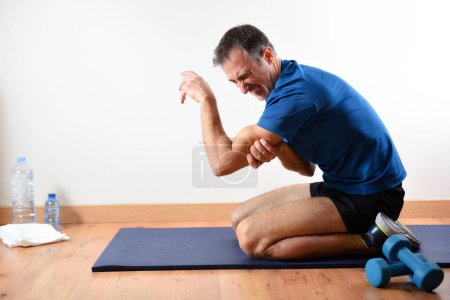 Hombre haciendo deportes en el interior quejándose de dolor de tríceps sentado en una alfombra. Vista lateral