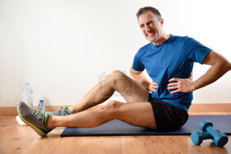 Photo for Man doing sports indoors complaining of ribs pain sitting on a mat. Side view - Royalty Free Image