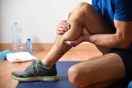 Foto de Detail of man doing sports with calf pain holding himself with his hand sitting on a mat. Side view - Imagen libre de derechos