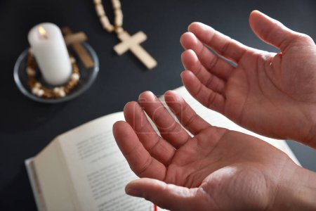 Foto de Cristiano rezando con las manos arriba con la mesa negra en el fondo con la cruz cristiana de la biblia y la vela encendida. - Imagen libre de derechos