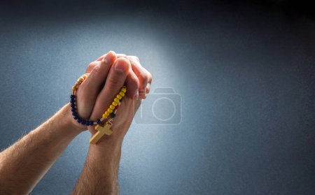 Christian praying with hands together with fingers interlocked with rosary beads hanging and isolated dark background. Top view.