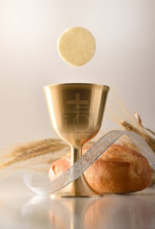 Recordatorio de la primera comunión con una copa de cáliz con una cruz grabada y una hostia sobre una mesa con una barra de pan y espigas de trigo. Composición vertical. Vista frontal.