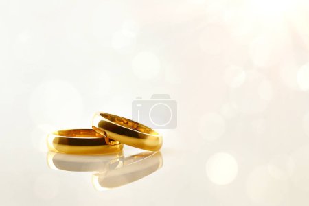 Detalle de dos anillos de oro uno encima del otro reflejado en una base pulida con fondo dorado con bokeh. Vista frontal.