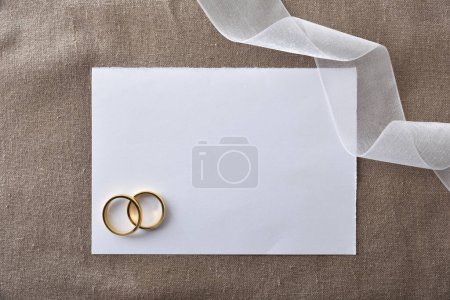 Téléchargez les photos : Annonce de mariage fond avec des anneaux d'or en papier découpé sur tissu brun et détail de ruban blanc. Vue du dessus. - en image libre de droit