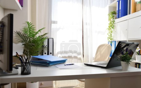 Téléchargez les photos : Bureau à domicile avec des meubles blancs avec des dossiers sur la table et les étagères et ordinateur portable sans personnes. - en image libre de droit