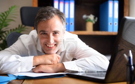 Empresario de negocios hombre de camisa blanca sonriendo y apoyado en una mesa de oficina con documentos y portátil y estantes con carpetas en el fondo