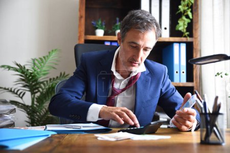 Photo for Stressed businessman doing financial calculations on a calculator on a wooden office table full of documents in an office with bookshelves in the background - Royalty Free Image