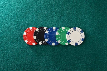 Casino-Hintergrund mit Details von fünf verschieden farbigen Plastikchips in einer Reihe auf einer grünen Filzspielmatte. Ansicht von oben.