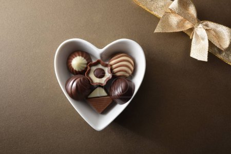 Foto de Bombones de chocolate surtidos en recipiente en forma de corazón de cerámica blanca sobre fondo de textura marrón con lazo de regalo de oro. Vista superior. - Imagen libre de derechos