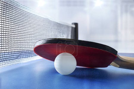 Tischtennispaddel ruht auf weißem Ball auf einem blauen Spieltisch neben dem Spielnetz mit Sportpavillon mit Beleuchtung im Hintergrund. Frontansicht.