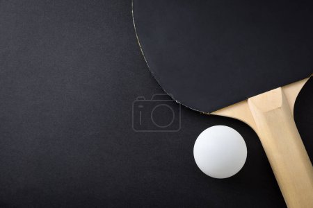 Detail of ping pong paddle with black rubber and white ball on black playing table. Top view.