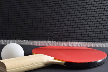 Ping pong set with paddle with red rubber and white ball on a black game table with net in the background. Front view.