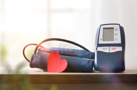 Blue digital blood pressure monitor for home on wooden table with cutout of red heart for blood pressure monitoring and window with curtains in the background. Front view.