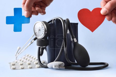 Concepto de monitorización de la presión arterial y la salud con monitor de presión arterial sobre fondo azul con las manos sosteniendo cruz médica azul y y corte cardíaco. Vista frontal.
