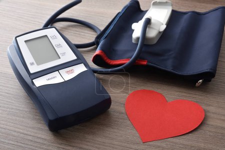 Blue digital blood pressure monitor for home on wooden table with cutout of red heart for blood pressure monitoring. Elevated view.