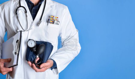 Detalle médico mostrando monitor de presión arterial vestido con bata blanca con carpeta de estetoscopio y medicamentos aislados sobre fondo azul. Vista frontal.