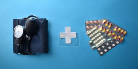 Medikamentenkonzept für Blutdruckprobleme mit Blutdruckmessgerät, Medikamentenblisterverpackung und weißem medizinischem Kreuzausschnitt auf blauem Hintergrund. Ansicht von oben.