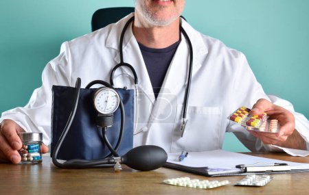 Médico que prescribe medicamentos para la presión arterial con varios paquetes de ampollas en las manos en una mesa de oficina de madera con la carpeta del monitor de presión arterial y más medicamentos. Vista frontal.