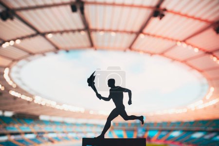 Foto de Silueta del atleta sosteniendo el relevo de la antorcha, fijada contra una pista moderna y un estadio del campo. Capturando el espíritu del evento de verano 2024 en París - Imagen libre de derechos