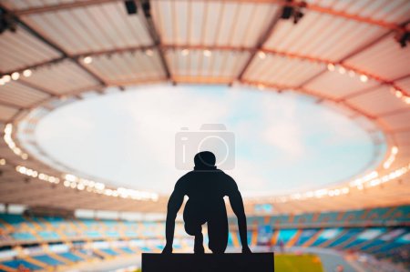 Photo for Silhouette of a Sprinter, Ready at the Starting Blocks, Against a Modern Sports Stadium Bathed in Beautiful Evening Light - Royalty Free Image