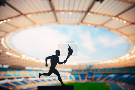 Foto de Silueta del atleta masculino que lleva el relevo de la antorcha, iluminando la pista moderna y el estadio de campo. Una cautivadora instantánea para el juego de verano 2024 en París. - Imagen libre de derechos