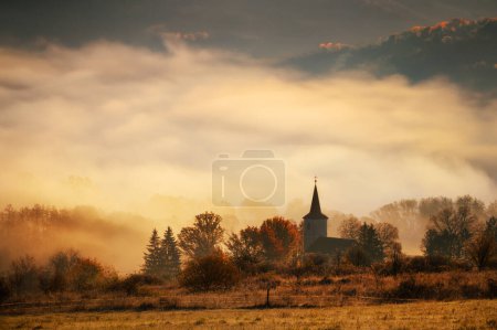 Foto de Cuadro con el motivo de la fe cristiana, la silueta de la iglesia cristiana, el sol de la mañana y la niebla en el fondo. Verano indio, colores anaranjados del amanecer - Imagen libre de derechos