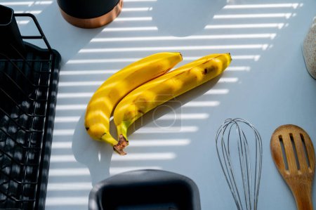 Téléchargez les photos : Banane sur le comptoir de la cuisine illuminée à l'ombre des stores. Lumière naturelle blanche, fond frit - en image libre de droit