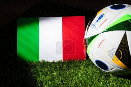 Foto de HAMBURG, GERMANY, APRIL 17, 2024: Italy national flag and official soccer ball of Euro 2024 football tournament in Germany placed on green grass. Black background, edit space - Imagen libre de derechos