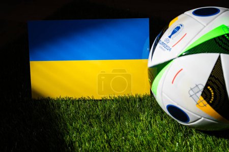 Photo for MUNICH, GERMANY, APRIL 17, 2024: Ukraine national flag and official soccer ball of Euro 2024 football tournament in Germany placed on green grass. Black background, edit space - Royalty Free Image