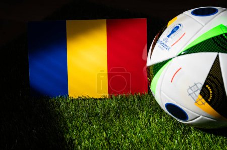 Photo for MUNICH, GERMANY, APRIL 17, 2024: Romania national flag and official soccer ball of Euro 2024 football tournament in Germany placed on green grass. Black background, edit space - Royalty Free Image