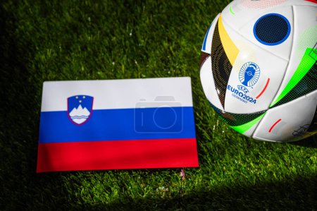 Photo for MUNICH, GERMANY, APRIL 17, 2024: Slovenia national flag and official soccer ball of Euro 2024 football tournament in Germany placed on green grass. Black background, edit space - Royalty Free Image