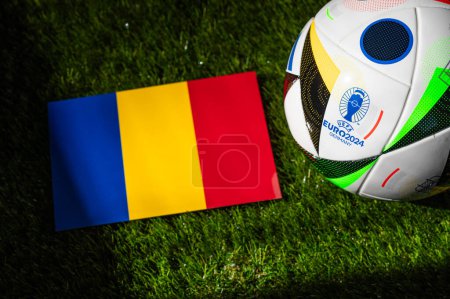 Foto de BERLÍN, ALEMANIA, 17 DE ABRIL DE 2024: Bandera nacional de Rumania y balón de fútbol oficial del torneo de fútbol Euro 2024 en Alemania colocado sobre hierba verde. Fondo negro, editar espacio - Imagen libre de derechos