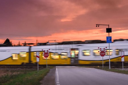 Foto de Un tren de pasajeros que pasa a través de un ferrocarril sin vigilancia que cruza bien marcado, cielo nocturno iluminado por el sol poniente. - Imagen libre de derechos